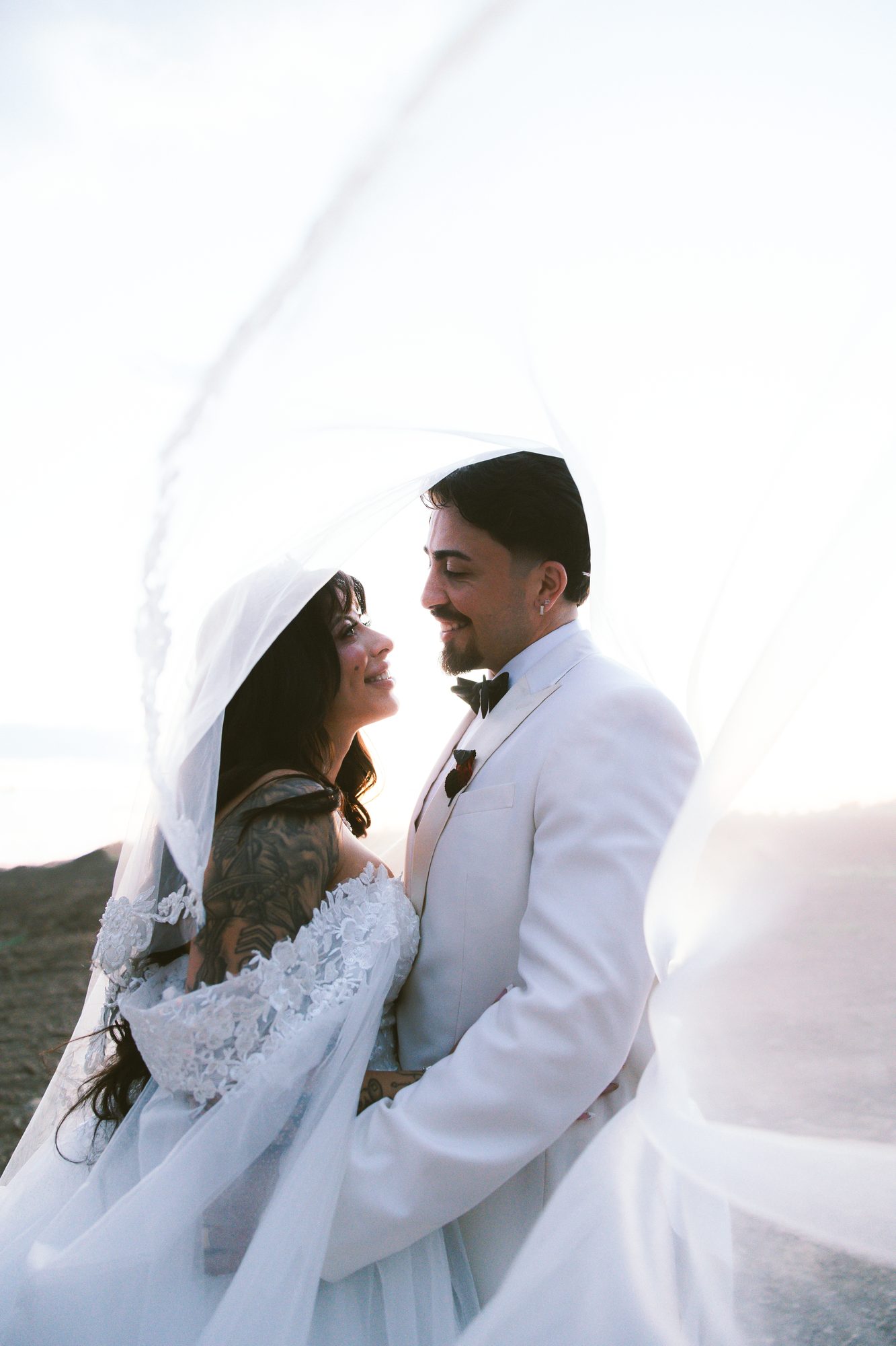 Bride and groom in white, her veil lifted by the wind at sunset