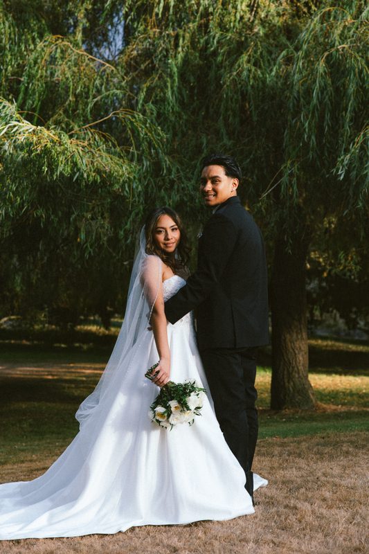 Couple under a willow tree, the bride holding white flowers